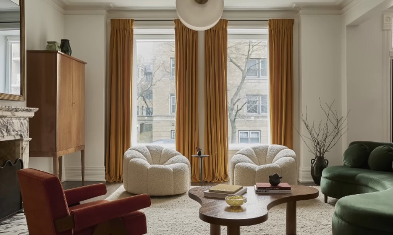 A stylish living room with white walls, a curved dark green velvet sofa, a burnt orange velvet armchair, and two white textured armchairs centered near a window. The windows are framed by long, deep gold/mustard yellow velvet curtains. A dark wood cabinet is on the left, and a unique, irregularly shaped wood coffee table sits on a light-colored area rug.