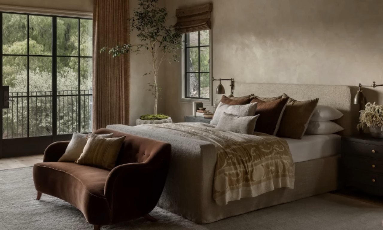A bedroom with warm, earthy tones featuring a large bed with a neutral upholstered headboard and layers of bedding. A small, curved, velvety brown settee sits at the foot of the bed, and a large rug anchors the space. A potted tree stands next to the bed near a glass door leading outside.