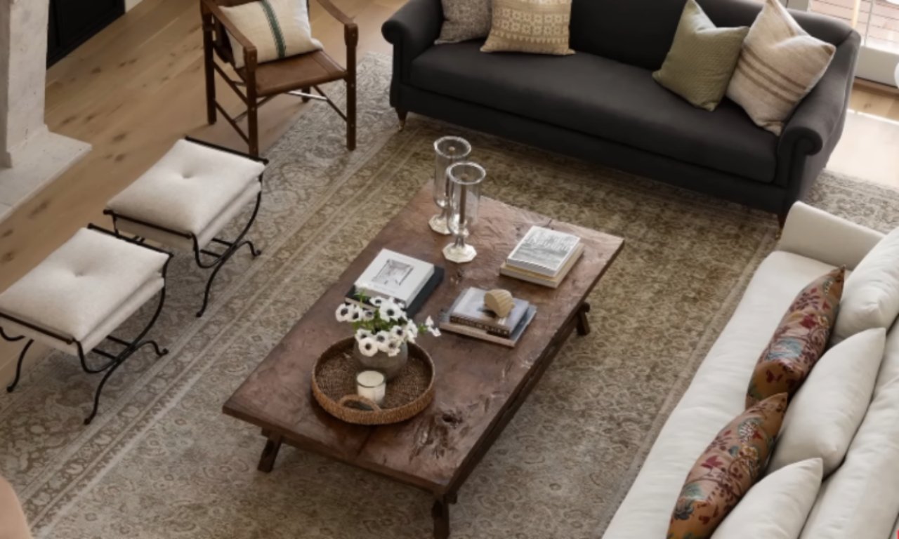 An overhead view of a living room showing a dark brown sofa facing a white sofa across a rustic, rectangular wooden coffee table. A vintage-style neutral-toned area rug with a subtle pattern covers the floor. The coffee table holds books, glass vases, and a wicker tray with flowers and a candle. Two white upholstered stools with dark metal bases are visible in the top left corner.