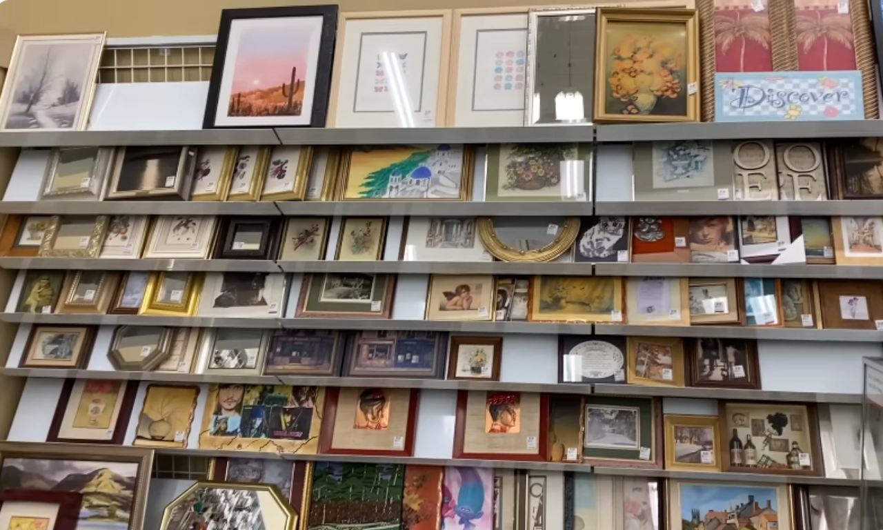 A floor-to-ceiling shelving unit fully stocked with a large and eclectic collection of framed wall art and pictures. The display includes various sizes, frames, and subjects, suggesting a retail or thrift store environment.