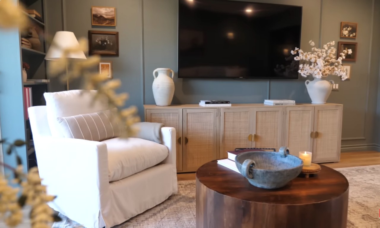 A cozy, stylish living room featuring a large, white slipcovered armchair in the foreground with a striped lumbar pillow. In the background, a large black TV is mounted above a light wood, woven-front media console. The walls are a dark sage green with paneling, and the decor includes a large white ceramic vase with branches, framed artwork, a floor lamp, and a round wooden coffee table.