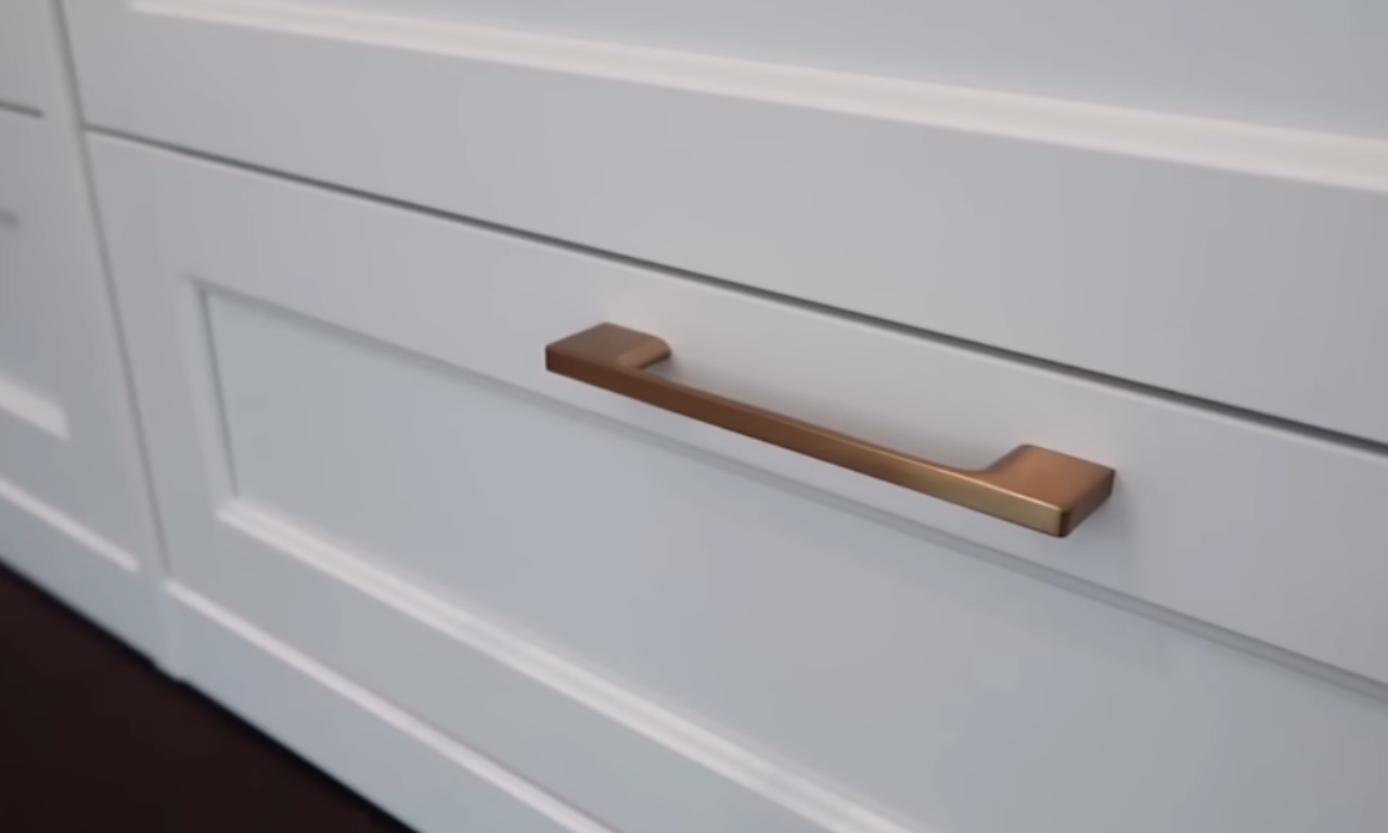 A close-up shot of a white kitchen or bathroom cabinet drawer featuring a sleek, brushed brass or gold-toned pull handle. The handle is a long, modern bar style.