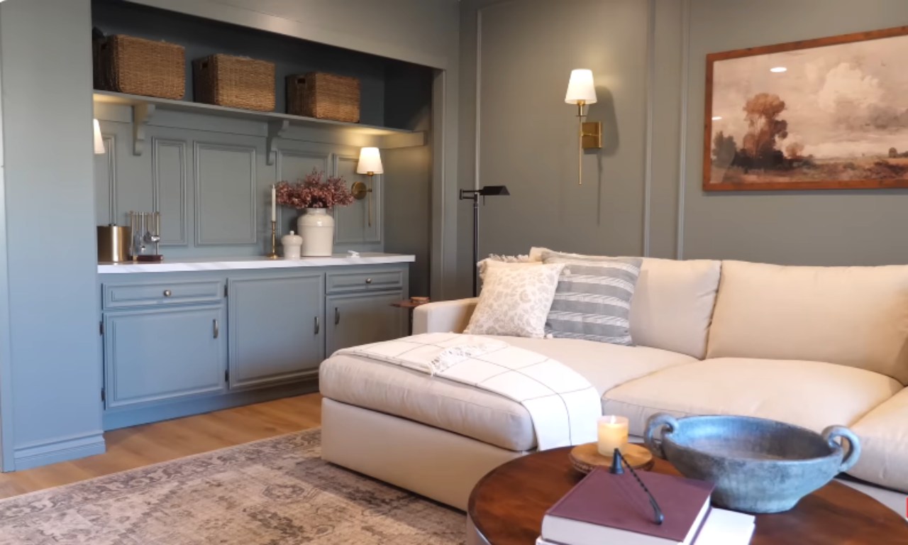 A close-up view of a comfortable beige sectional sofa on a patterned area rug. To the left is a custom built-in cabinet and counter area painted a dusty light blue, featuring woven baskets on a shelf above. The walls are a dark sage green with wainscoting. A round, dark wood coffee table in the foreground holds a large decorative bowl and a candle. Sconces with brass accents are mounted on the wall above the sofa and on the built-in.