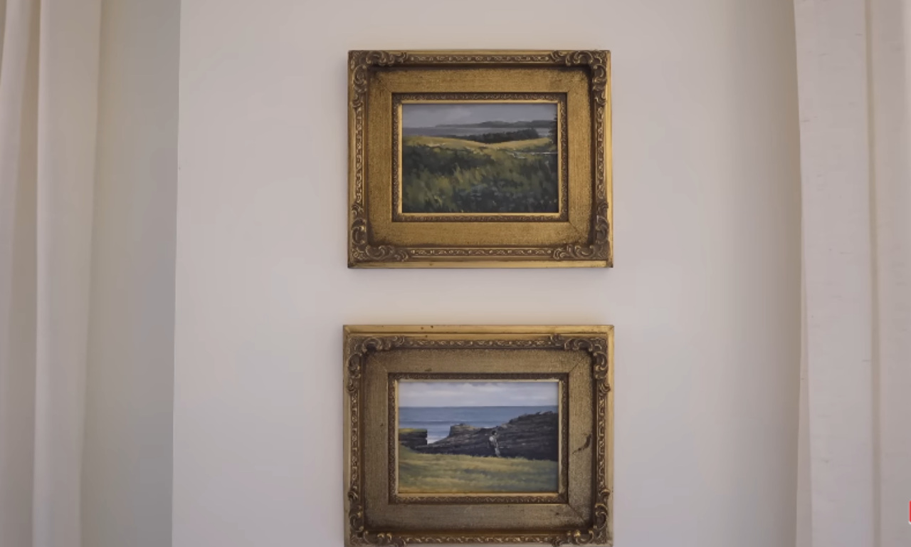 wo identical square oil paintings of landscape scenes, one above the other, hung on a white wall. Both paintings are in ornate, distressed gold-colored frames. The top painting depicts a grassy, hilly field leading to a body of water under a gray sky. The bottom painting shows a similar grassy foreground, a rocky coast, and a choppy blue-gray sea.