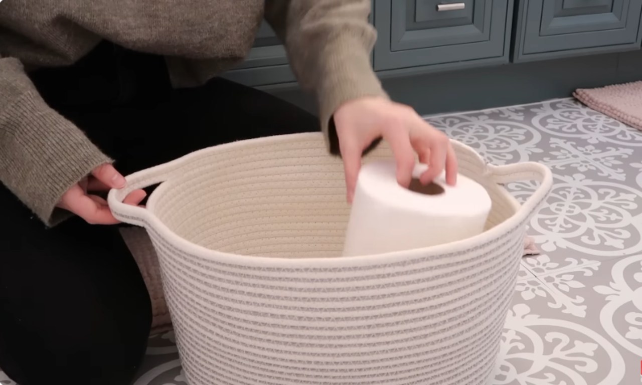 A person, dressed in dark clothing, is placing a roll of toilet paper into a large, cream-colored, woven rope storage basket with gray accents. The basket sits on a gray and white patterned floor in front of a teal/blue vanity.