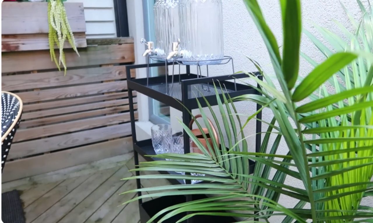 A close-up shot of a black metal bar cart holding two large glass drink dispensers with spigots, and a couple of clear glasses. A copper watering can rests on a lower shelf. Green palm fronds are visible in the foreground.