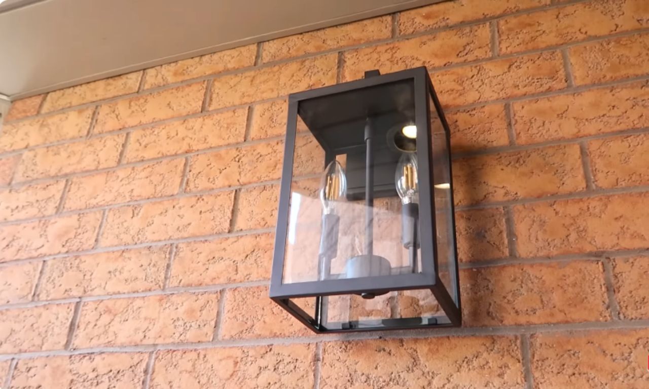 A close-up view of a modern, black-framed outdoor wall lantern with clear glass panels mounted on a red brick wall. The lantern is lit and holds two decorative Edison-style LED bulbs.