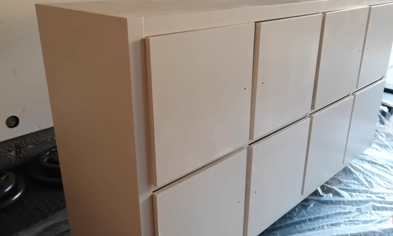 A large, pale pinkish-beige dresser or cabinet unit is shown partially assembled, possibly in a garage. It has a smooth top and six rectangular drawer or cabinet fronts (three on top, three on the bottom) that are currently smooth and unadorned, waiting for hardware or further modification. The unit is sitting on a plastic drop cloth.