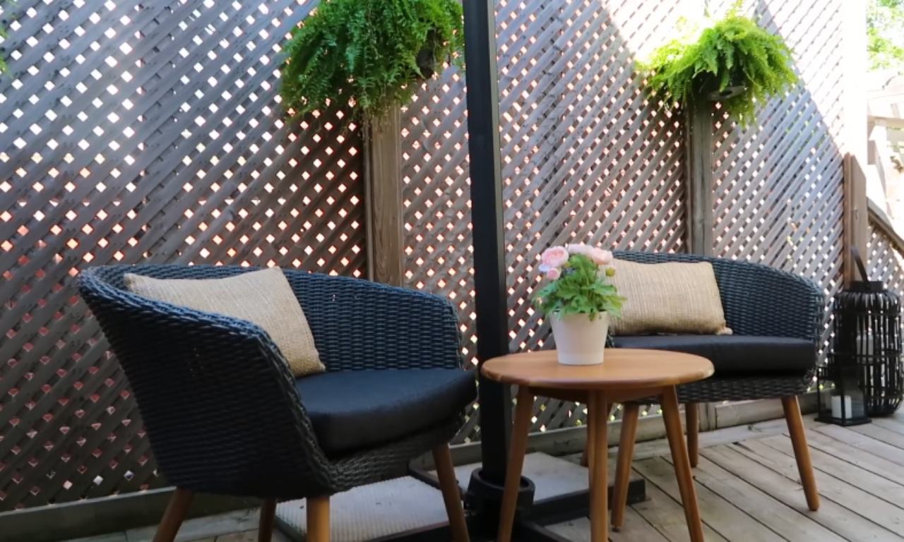 A cozy outdoor seating area featuring two black woven armchairs with tan pillows, separated by a small, round wooden side table with a potted plant. The area is backed by a lattice fence with hanging ferns, all resting on a wooden deck.