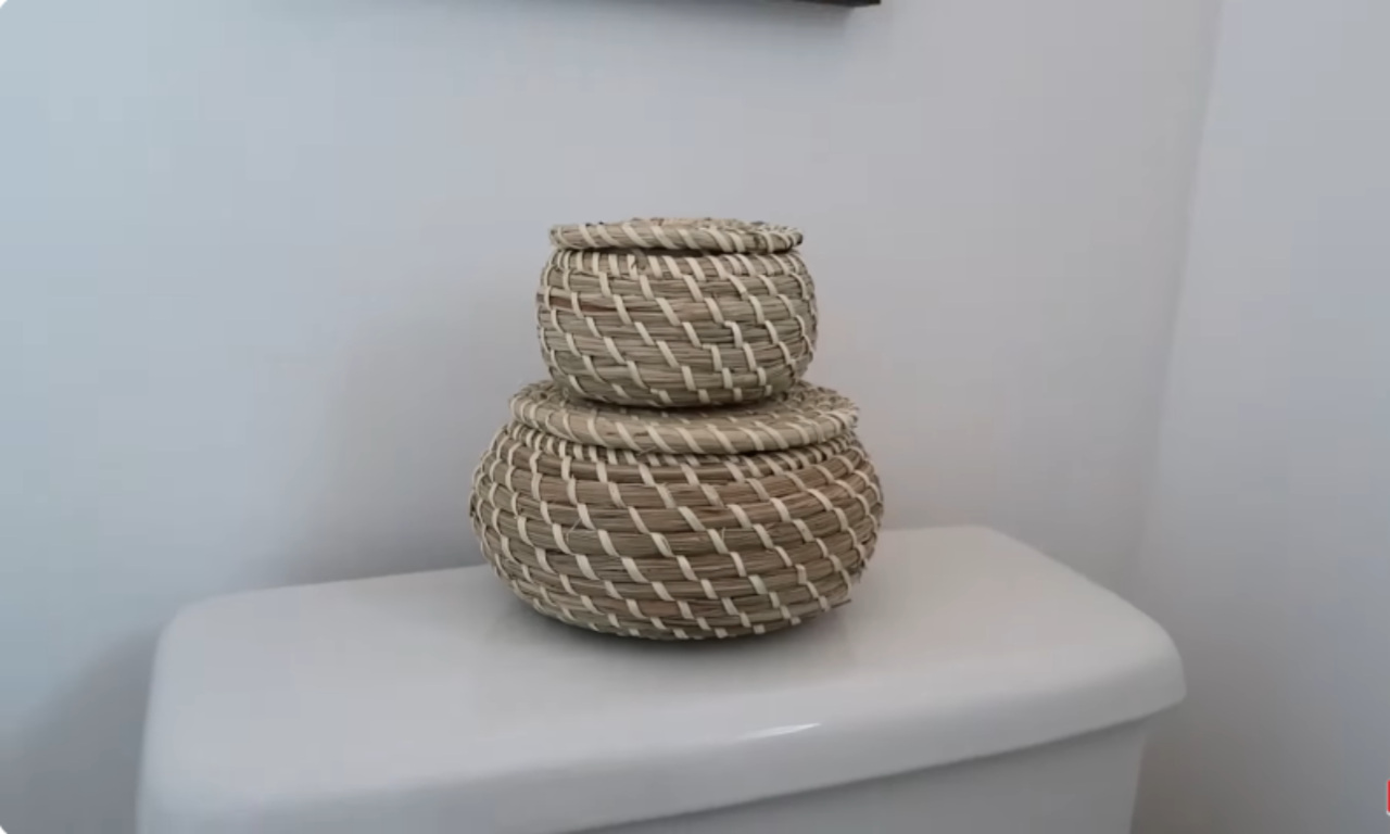 Two small, woven natural fiber baskets with lids are stacked on top of a white toilet tank. The baskets have a light brown and off-white woven pattern. The wall behind the toilet is plain white.