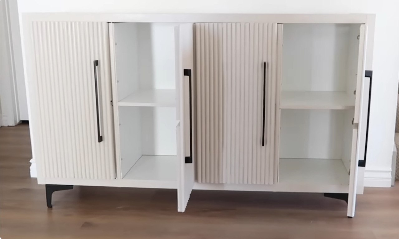 A finished, pale off-white cabinet with four doors open, revealing two storage sections with shelves inside. The two outer doors and the two inner doors all feature a vertical fluted texture. Each door has a long, modern, black bar pull handle. The cabinet is elevated on short, black, angled furniture legs. This appears to be the final result of a furniture hack project.
