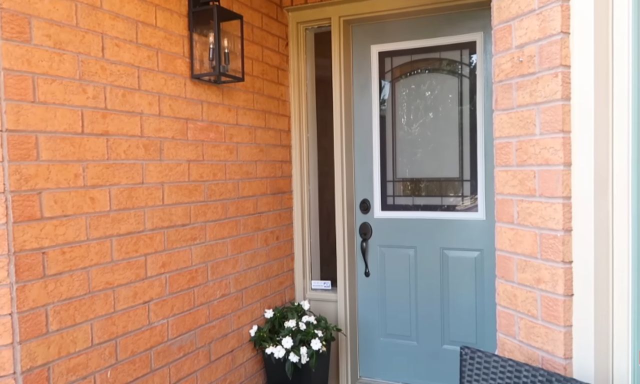 An outdoor scene showing a light blue or teal front door with a window featuring decorative glass. To the left, a modern black outdoor lantern is mounted on the red brick wall, and a black planter with white flowers sits near the door.