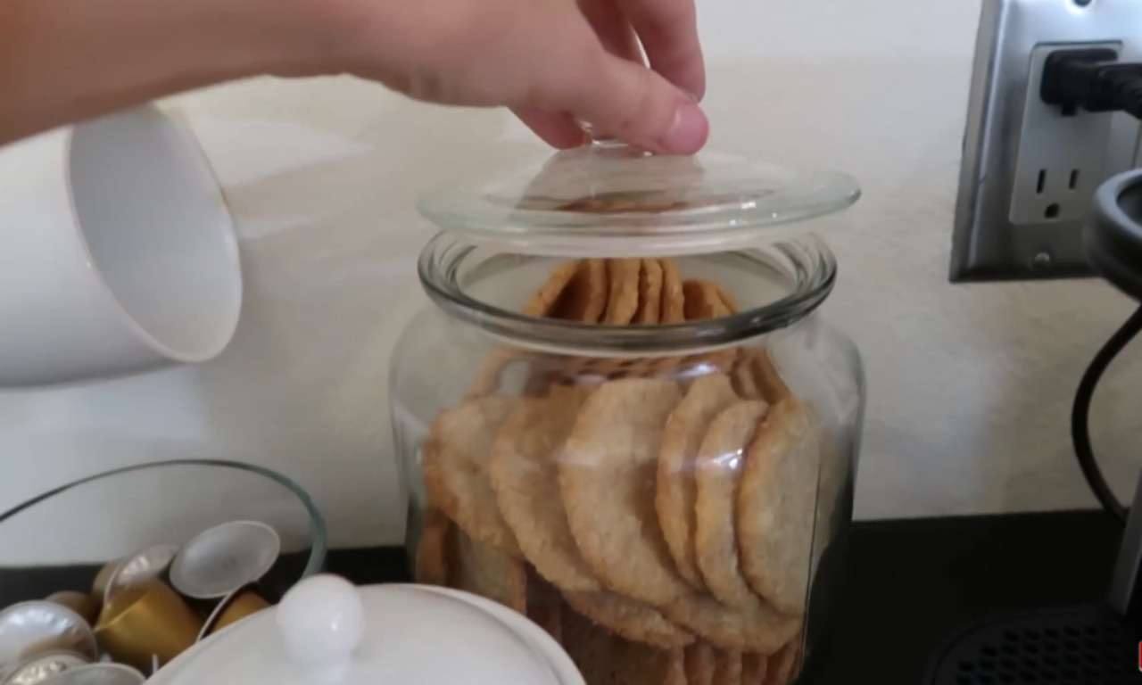 A close-up of a hand lifting the glass lid off a large, clear glass jar filled with golden-brown cookies or crackers, next to a white ceramic sugar bowl and a bowl of metallic coffee pods. An electrical outlet is visible in the background.