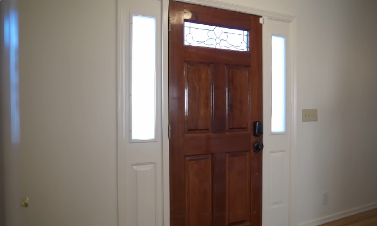 A close-up, interior view of a home's front entry door. The door is a rich, warm wood tone with four panels and a decorative stained glass window insert at the top. It is flanked on both sides by white sidelights and is set into a plain, white wall. A smart lock is visible on the door.