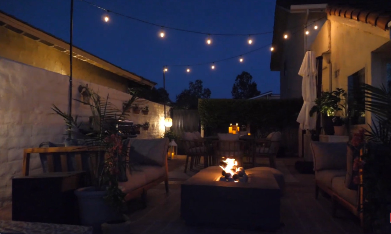 A nighttime patio scene with string lights overhead, a lit gas fire pit, and candlelight on a dining table.