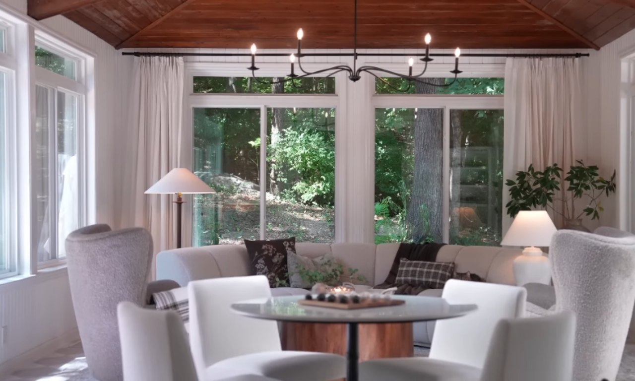 A sunroom with a vaulted wooden plank ceiling and a large window wall overlooking a lush wooded area. The main seating area includes a cream-colored sectional sofa flanked by two textured, curved armchairs. A small, modern glass-top dining table with four white chairs is in the foreground. The room is decorated with long white curtains, a black metal chandelier, and white lamps on side tables.