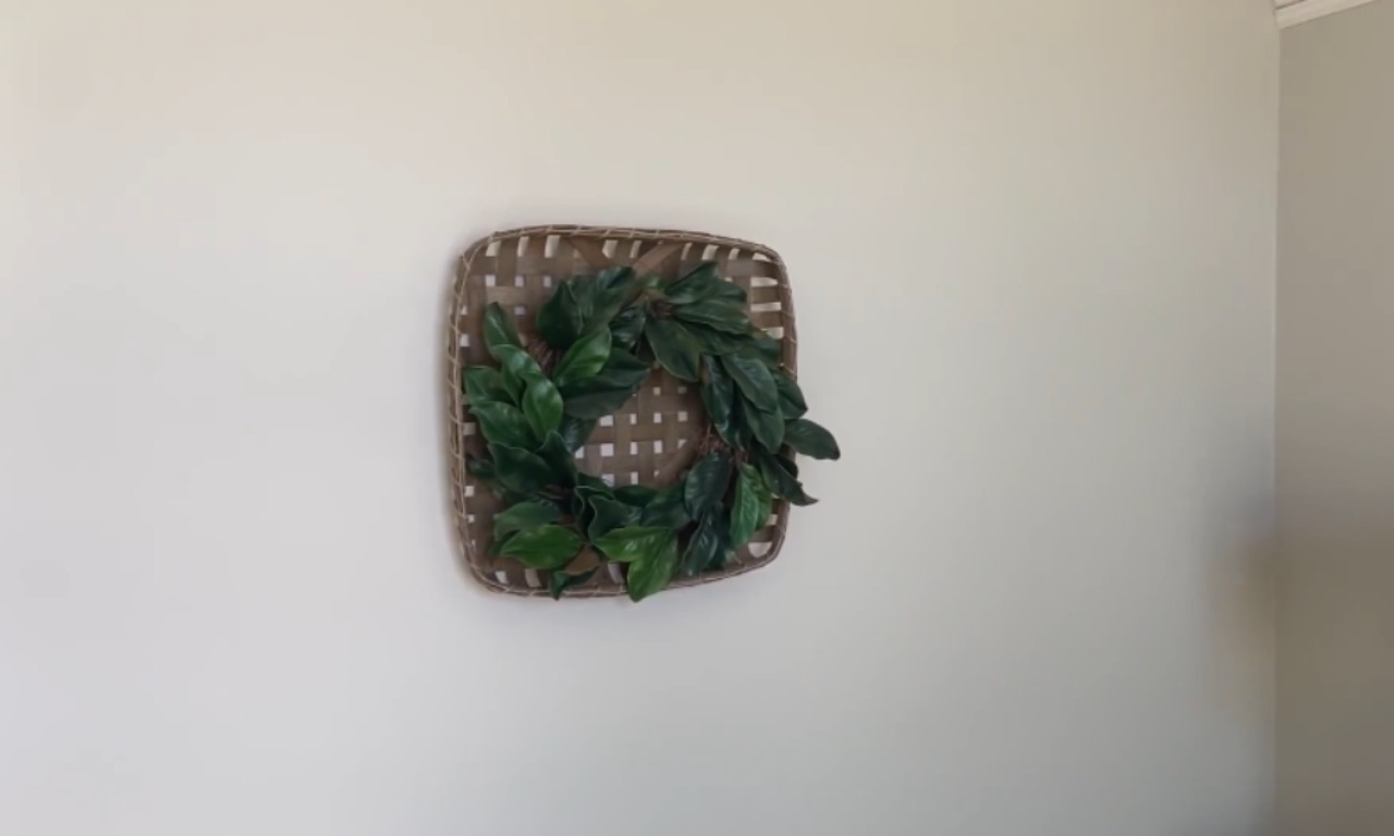close-up of a light-colored wall featuring a square, woven tobacco basket decorated with a small, simple wreath of deep green, leafy foliage.