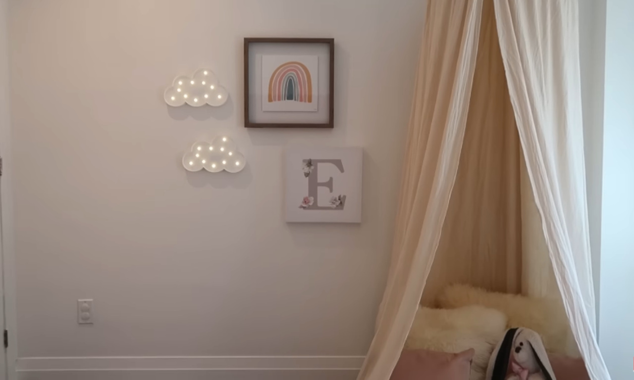 A close-up view of a child's bedroom wall painted a creamy white. Decor includes two light-up cloud shapes, a framed picture of a muted rainbow, and a framed canvas with the letter 'E' decorated with floral and leaf motifs. To the right, a sheer, light beige canopy hangs over a corner, revealing a peek of soft bedding and a stuffed bunny toy.