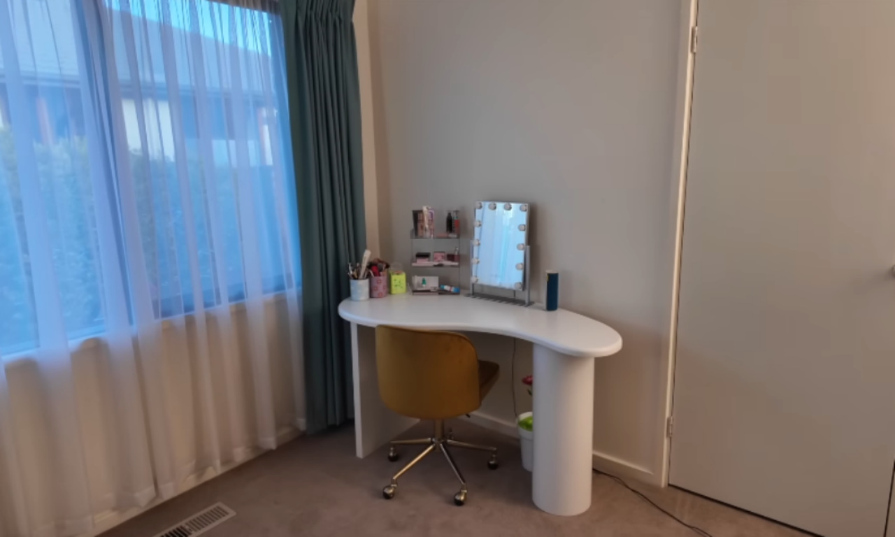 A bright, corner desk with a white, curved top is positioned by a large window with sheer, pale-teal curtains. The desk has a small vanity mirror with lights, some makeup, and brushes. A mustard-yellow upholstered chair is tucked beneath the desk. The walls are a light beige/gray.