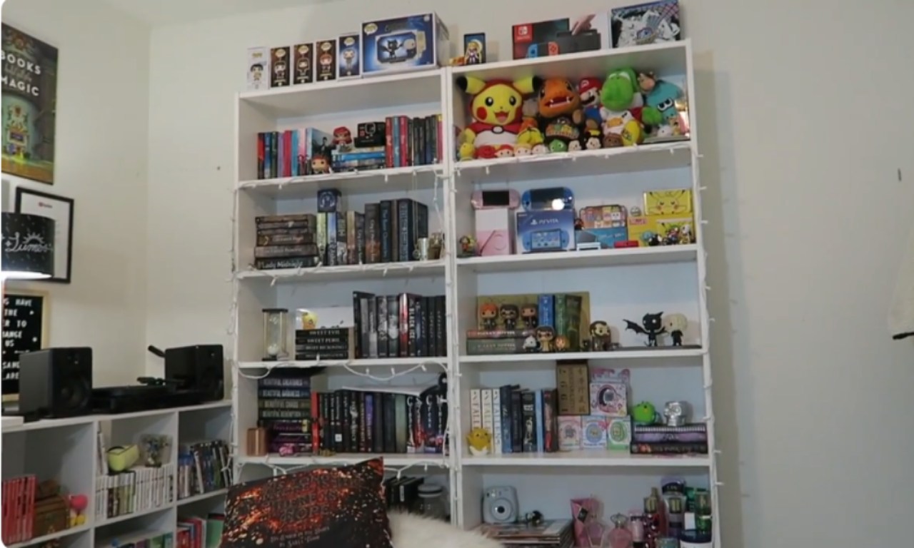 A tall, white double-shelving unit packed with books, video games, consoles, Funko Pop figures, and various stuffed animals, including a large Pikachu. The shelves are brightly lit, suggesting a bedroom or living space.