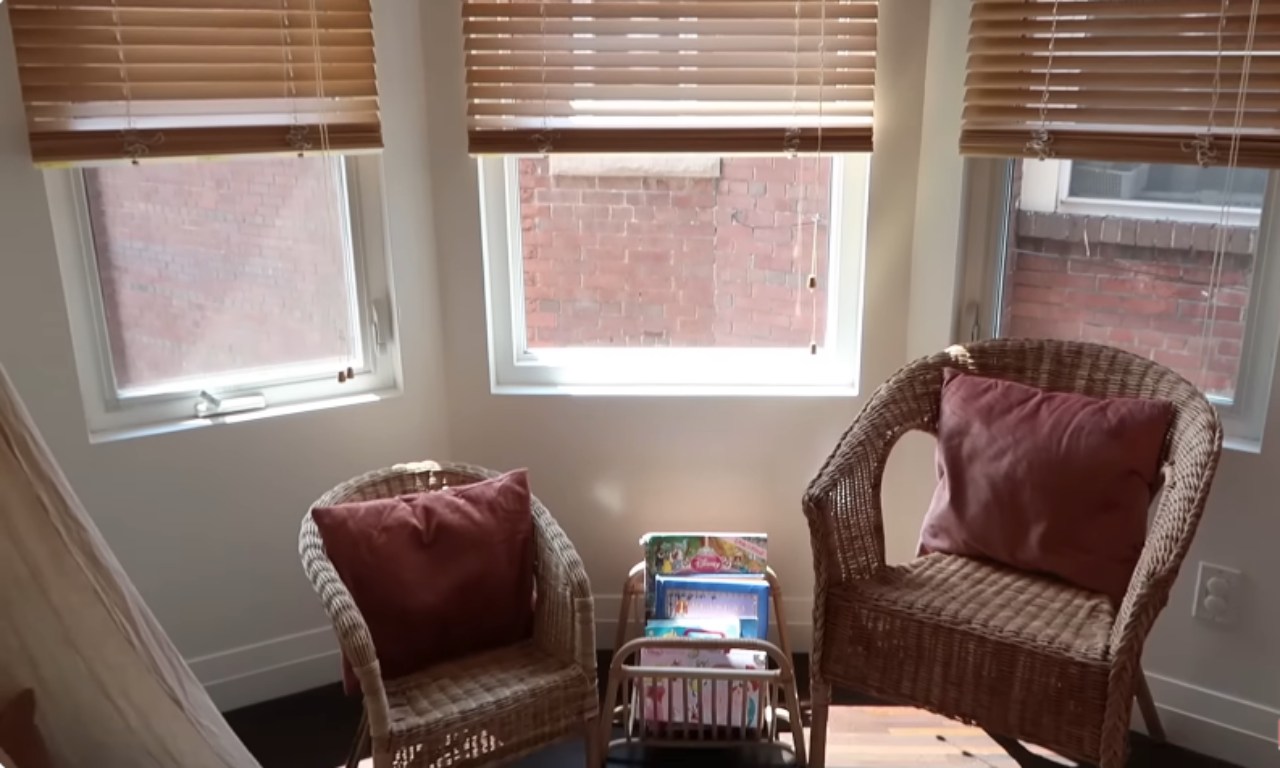 A cozy reading nook in a child's bedroom with two light brown wicker chairs facing each other, each topped with a terracotta-colored cushion. Between the chairs sits a small, wooden, rolling bookshelf filled with children's books. The area is brightly lit by three large windows dressed with light brown wooden blinds.