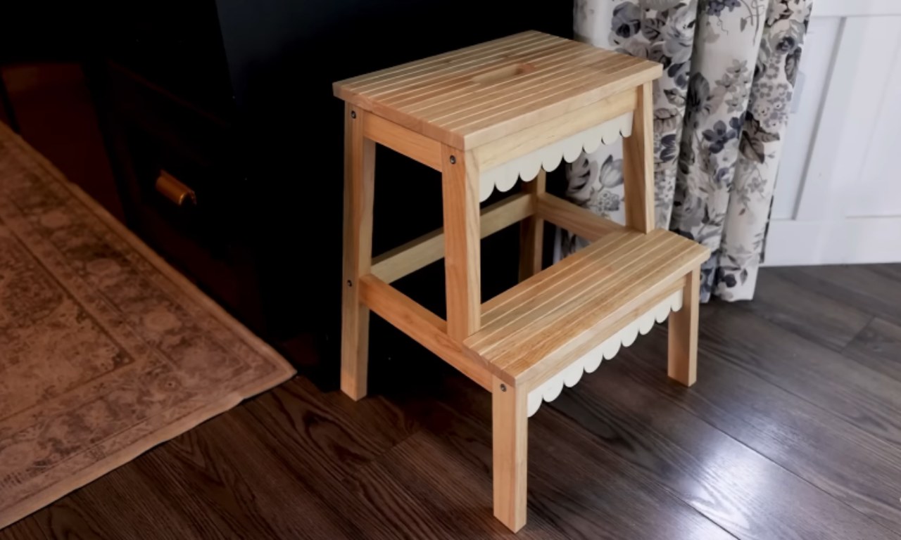 A light wood IKEA step stool, likely the BEKVÄM, that has been customized with decorative scalloped white trim added beneath the edges of both steps. It sits on a dark wood floor next to a black dresser and a patterned curtain.