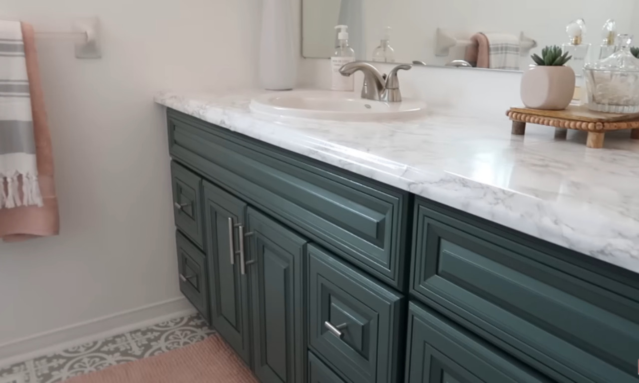A close-up of a modernized bathroom vanity. The cabinet is painted a deep teal or hunter green with silver hardware. The countertop is a white marble-look laminate or contact paper. A white ceramic sink, silver faucet, and a small potted succulent are visible on the counter. A pink and gray towel hangs on a nearby rack, and a pale pink rug is on a gray and white patterned floor.