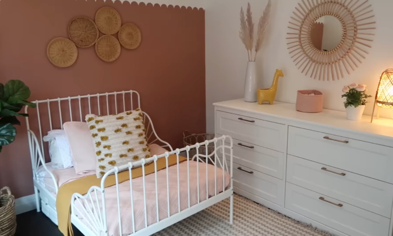 A toddler's bedroom featuring a white metal bed with railings and soft pink and mustard yellow bedding. The wall behind the bed is painted a dusty rose or terracotta color with a decorative scalloped trim at the ceiling. Above the headboard, there is a gallery arrangement of five woven wicker plates. To the right, a white dresser is topped with a large circular rattan sunburst mirror and a few decorative items, including a yellow ceramic giraffe, pampas grass in a white vase, and a small woven lamp.