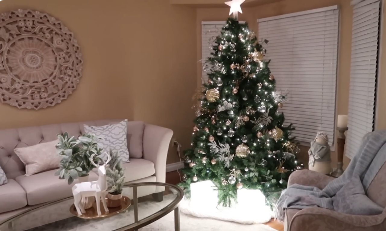 A beautifully decorated Christmas tree, lit with white lights and adorned with gold and white ornaments, stands in the corner of a living room. The base of the tree is covered with a white, soft tree skirt. To the left is a tufted light-colored sofa with patterned throw pillows and a glass coffee table holding a gold tray with small deer figurines and faux greenery. To the right is an armchair with a light blue throw blanket. A large, circular, carved wooden wall hanging is visible on the tan wall.
