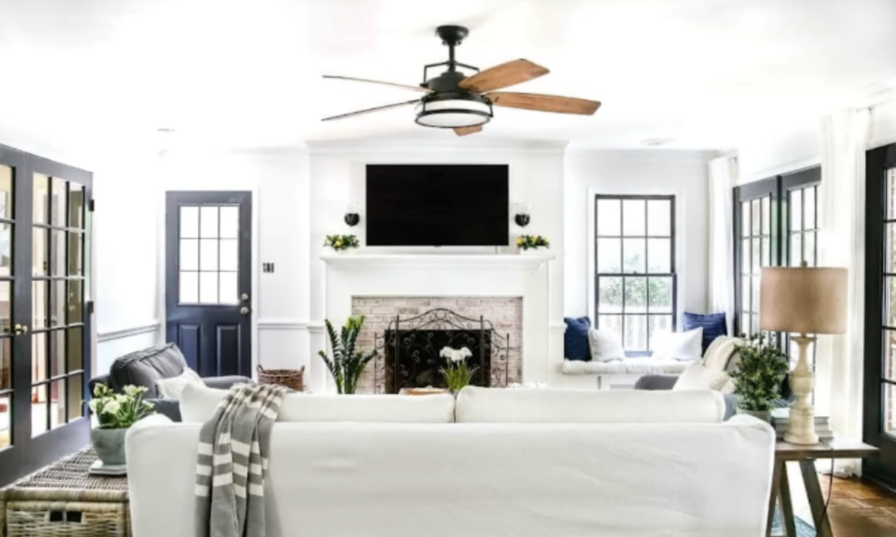 A bright, cozy living room. A white slipcovered sofa faces a fireplace with a white mantel and a mounted television above it. The room has light walls, dark wood accents, including a ceiling fan and french doors with black frames.