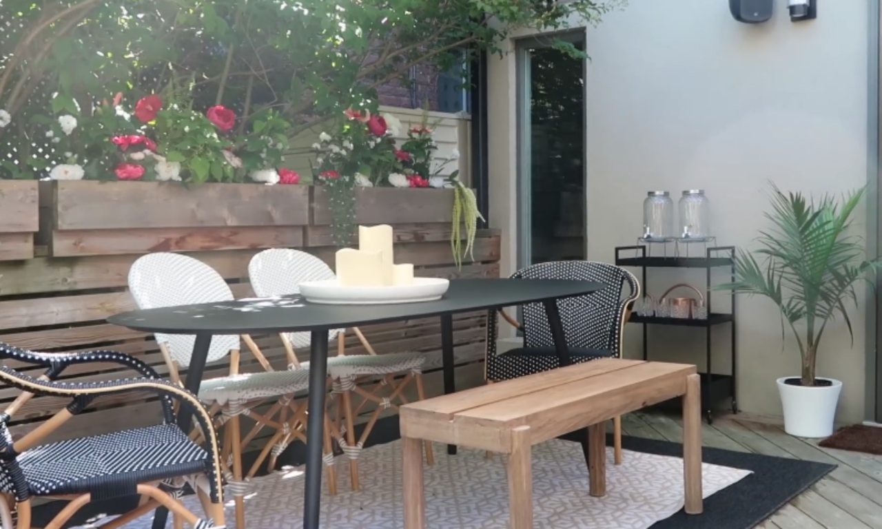 A stylish small patio features a black oval table, a wooden bench, and black and white woven chairs on a patterned outdoor rug. A wooden planter box with pink and white roses is visible along the back fence. A black rolling cart with glass drink dispensers and a potted palm stands to the right.