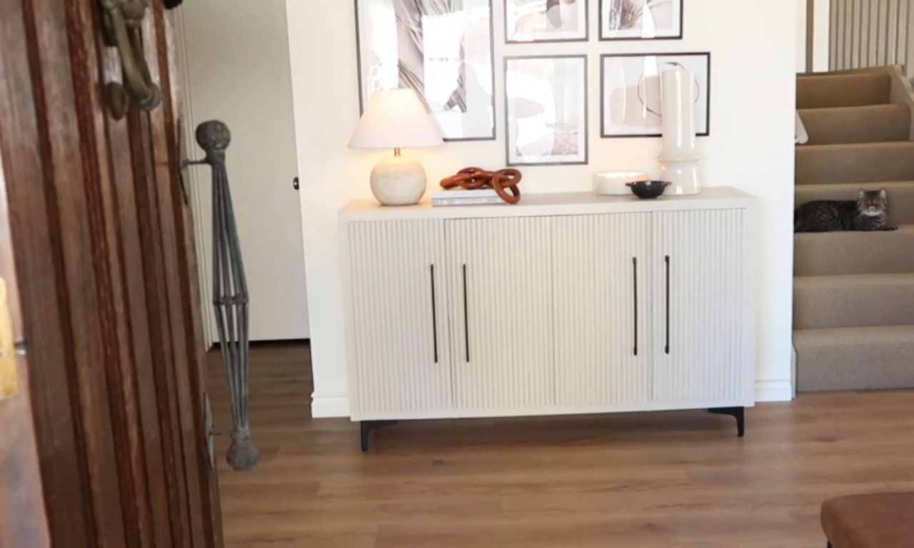 A modern entryway featuring a light-colored, textured four-door console cabinet with thin black vertical handles and black legs. On the console are a marble-base lamp, a coiled brown decorative object, a small white bowl, and a tall white sculptural vase. Above the cabinet is a gallery wall of framed neutral-toned art prints. To the left is a partially open dark wood door. To the right, a cat sits on the carpeted stairs. The flooring is a warm-toned wood laminate.
