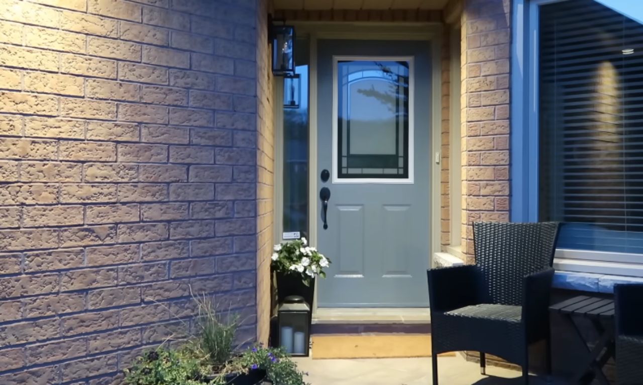 An evening view of a well-lit front entrance. The light blue or gray front door is visible, along with the adjacent red brick wall. A modern black lantern illuminates the area, which includes a doormat, a planter with foliage, and a small outdoor seating arrangement with black wicker chairs.