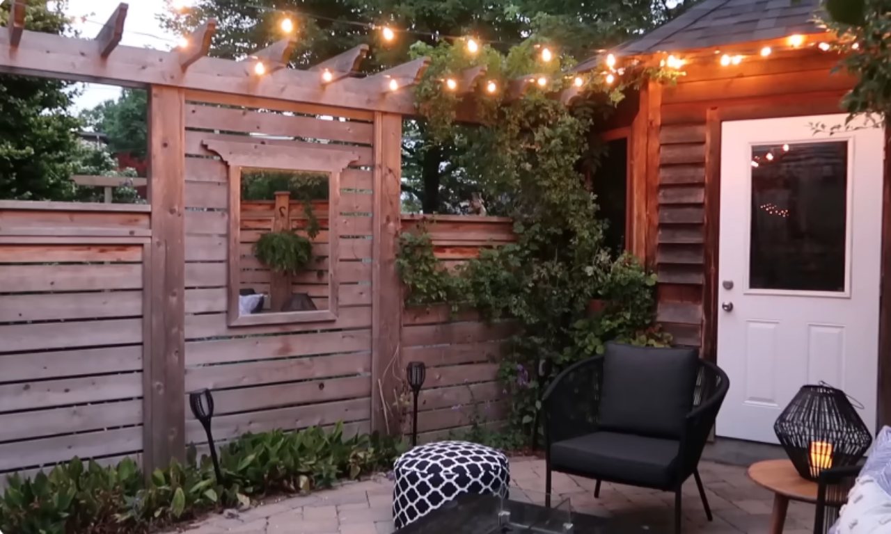 A cozy outdoor patio or backyard seating area at dusk. The space is defined by a wooden privacy fence and pergola structure accented with string lights overhead. A dark, woven outdoor chair and a black and white patterned ottoman are visible on a paved stone patio. The wooden fence has a decorative rectangular opening, possibly holding a mirror or a plant. The area is bordered by lush greenery.