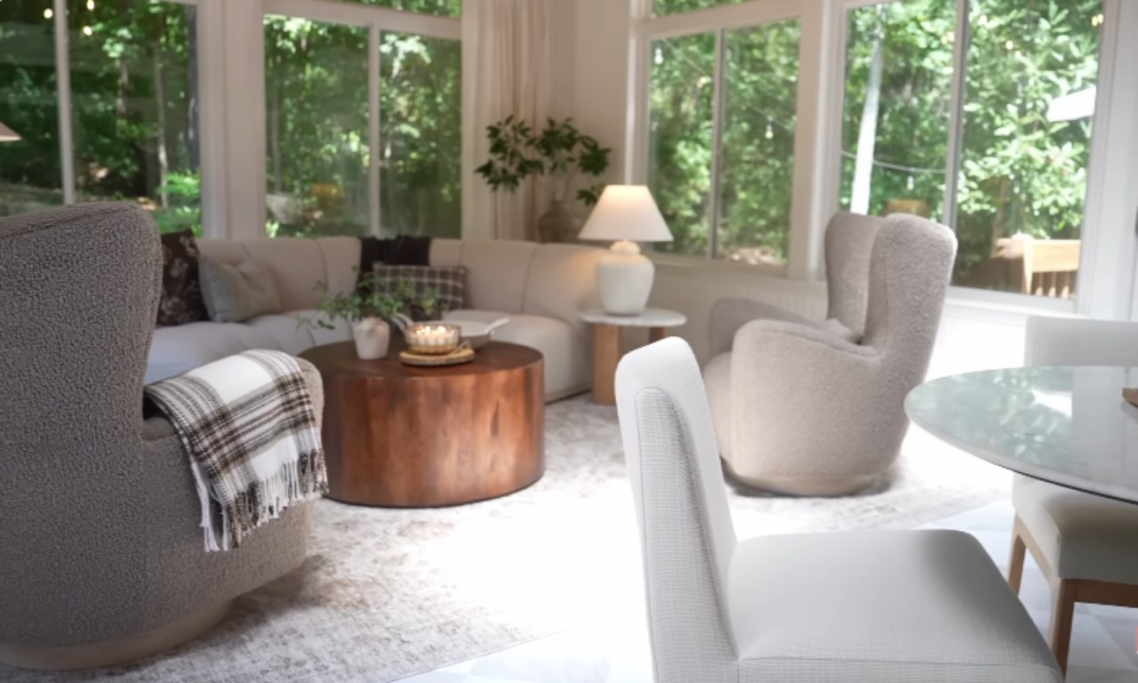 A bright, cozy sunroom with large windows overlooking a wooded area. The room features a cream-colored sectional sofa, two textured, curved armchairs, a large round wooden coffee table, and a small round side table with a white lamp. A plaid throw blanket is draped over one of the armchairs. White dining chairs and a glass-top table are visible in the foreground to the right.