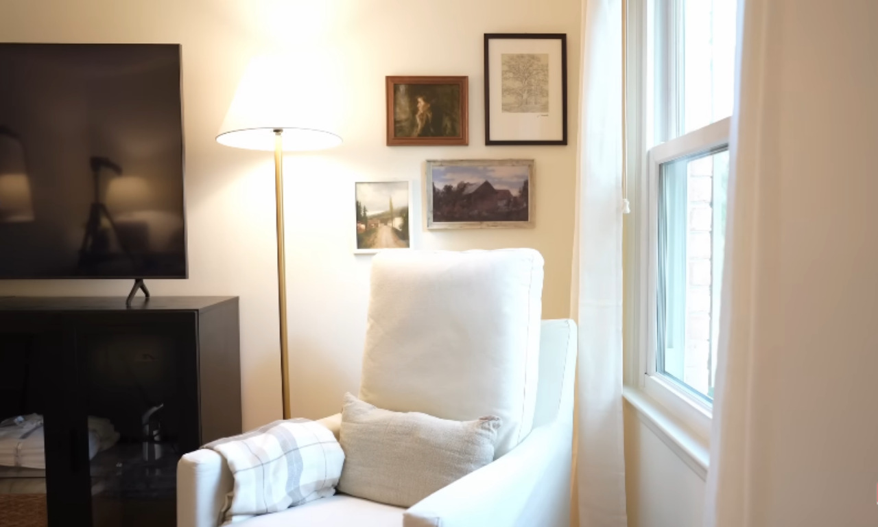 A small gallery wall grouping of four framed art pieces arranged above a white armchair in a room corner. To the right is a window with white sheer curtains. A tall brass floor lamp with a large white cone shade stands next to the chair, and a large black TV and console are visible on the left. The art pieces include a portrait, a landscape, and a botanical print.