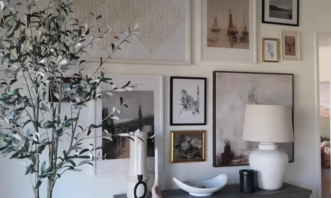 A styled home decor vignette featuring a large, faux olive tree in the foreground. In the background is a gallery wall with various framed art pieces, including a large abstract textile piece, a seascape, a monochrome city sketch, and an oil painting of flowers. A white ceramic table lamp with a linen shade sits on a dark gray console table, next to a decorative bowl and a tall candle. The overall style is modern and minimalist with a touch of classic art.
