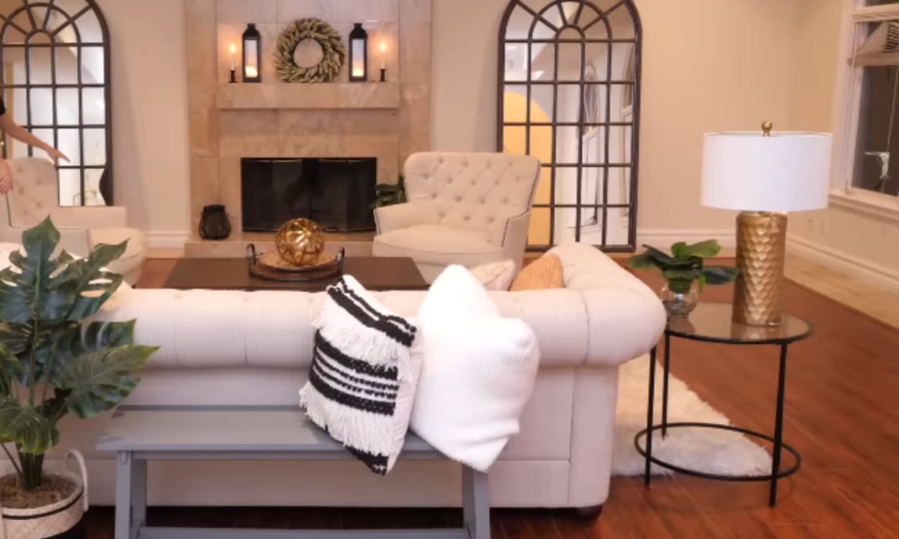 A formal living room with a fireplace as the focal point. The seating arrangement includes a cream tufted sofa facing the camera, with a small gray bench behind it. A large houseplant and a gold-based table lamp on a glass side table are visible on the right, and the fireplace wall is flanked by two tall, arched mirrors.