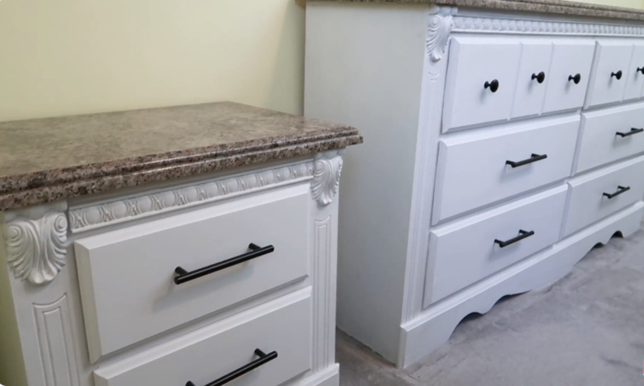 A side-by-side shot of a large white, chalk-painted dresser and a smaller matching nightstand. Both pieces have decorative carving, black modern bar-style drawer pulls, and a brown, faux-granite top.