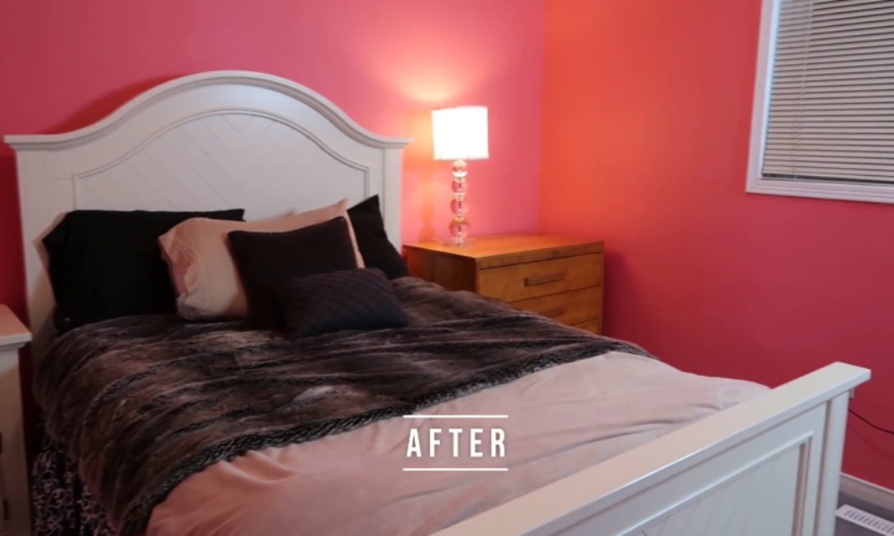 An "AFTER" shot of a neatly made bed with a white wooden headboard and a dark grey and light pink comforter. The wall behind the bed is painted a bright, vibrant pink, and a wooden nightstand with a clear glass lamp sits to the side. The room is clean and organized.