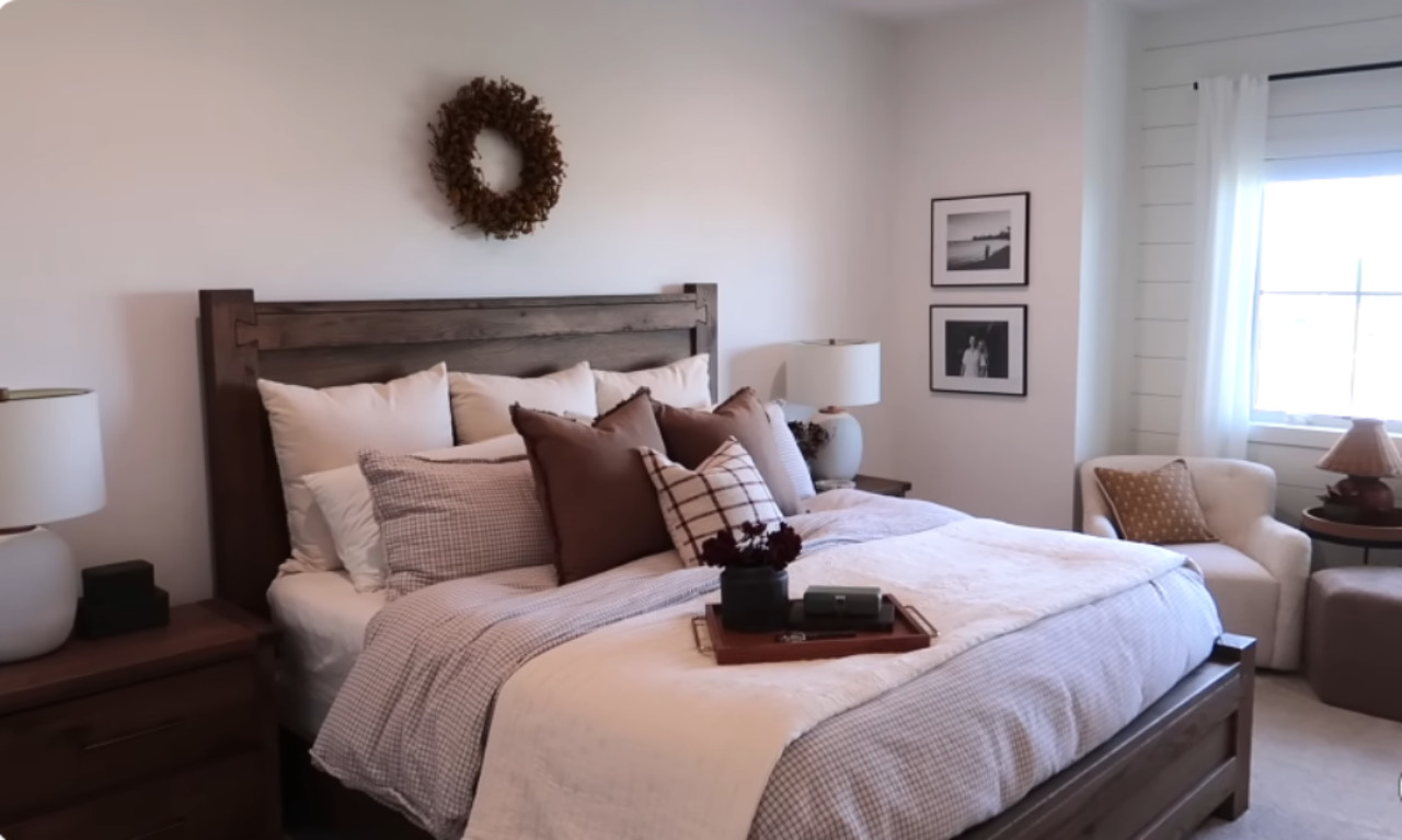 A decorated bedroom featuring a large dark wood bed frame with a layered bedspread in white, muted pink, and brown tones. The bed is adorned with numerous pillows in solid white, light brown, and checkered patterns. Above the headboard, a woven wreath is hung, and to the right, two framed black-and-white photos are on the wall. A nightstand with a lamp is on the left, and a small armchair sits in the far-right corner.