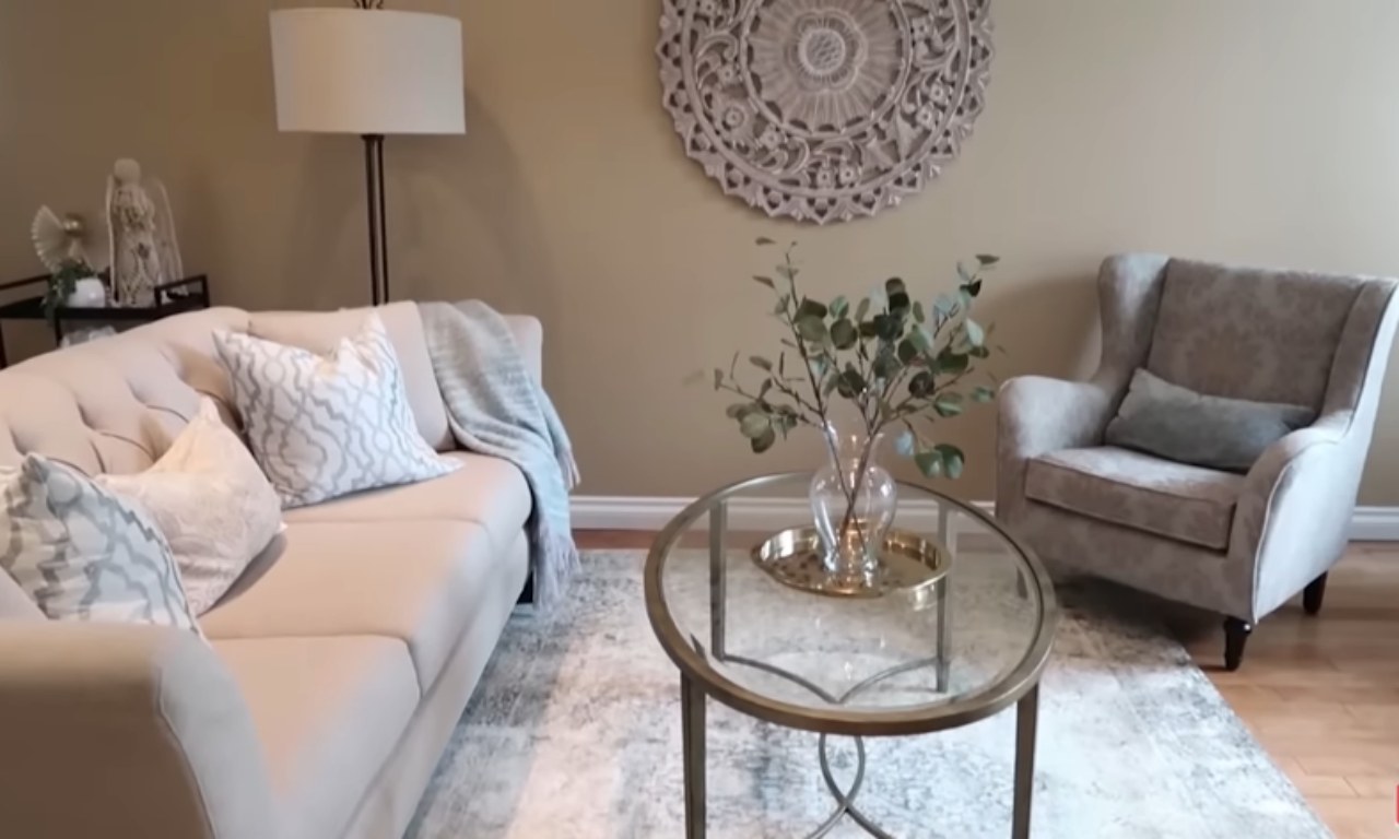 A well-lit, cozy living room featuring a neutral color palette. A light beige tufted sofa is on the left, draped with a light blue and white throw blanket and decorated with patterned pillows. In the center, a round glass coffee table with a gold metal base holds a clear vase with green eucalyptus branches and a small gold tray. To the right is a tall, upholstered wingback armchair. The tan wall behind the furniture is decorated with a large, intricately carved, round wooden medallion and a floor lamp stands near the sofa.