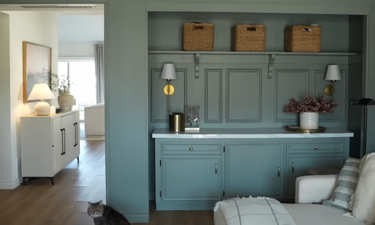 A cozy living room with light wood floors and a custom built-in cabinet and shelving unit painted a sage or dusty green color. The built-in features wainscoting-style paneling, a white marble countertop, and shelves topped with three large woven baskets. Two gold and white wall sconces are mounted on the built-in. To the left, a white credenza with a white lamp is visible in a brightly lit adjoining room. A cat is sitting on the floor in the foreground.