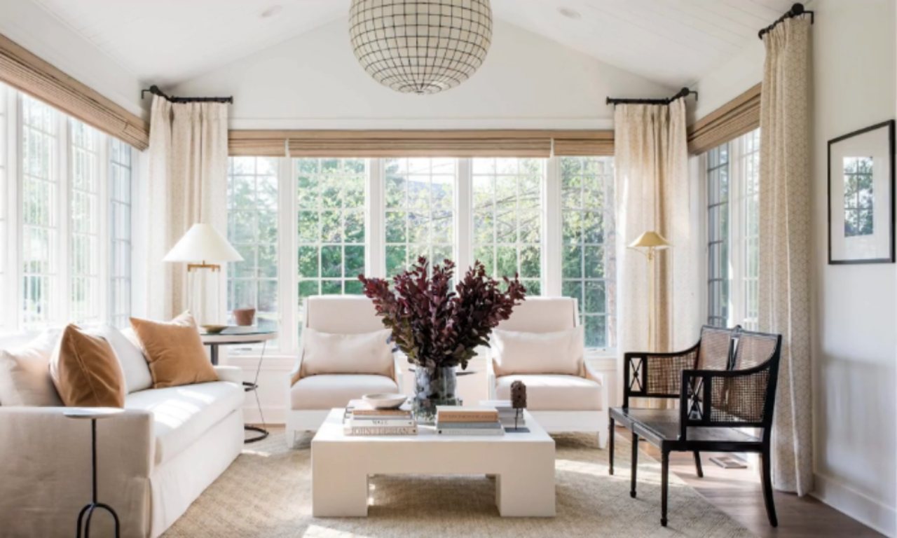 A bright, sunlit living room with a vaulted ceiling, featuring a white sofa and two light-colored armchairs centered around a square white coffee table. A large vase of deep red foliage sits on the table. The room has tall windows on three walls, covered by light-colored drapes and woven shades. A large, spherical pendant light fixture hangs from the ceiling. A dark wood and cane chair is to the right.