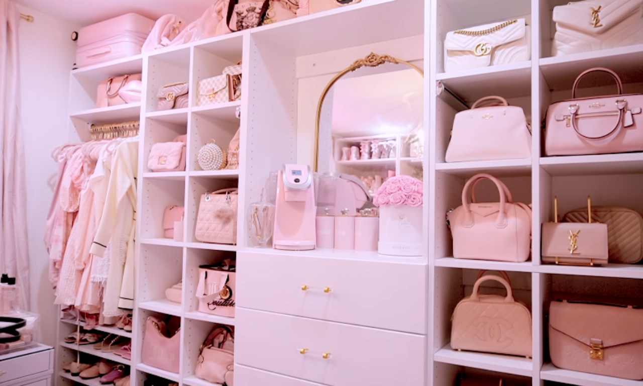 A highly organized, custom walk-in closet with white shelving and a predominantly pink theme. The shelves are filled with light pink clothing, designer handbags, and accessories. There is a central mirrored section with drawers and a small pink beverage maker.