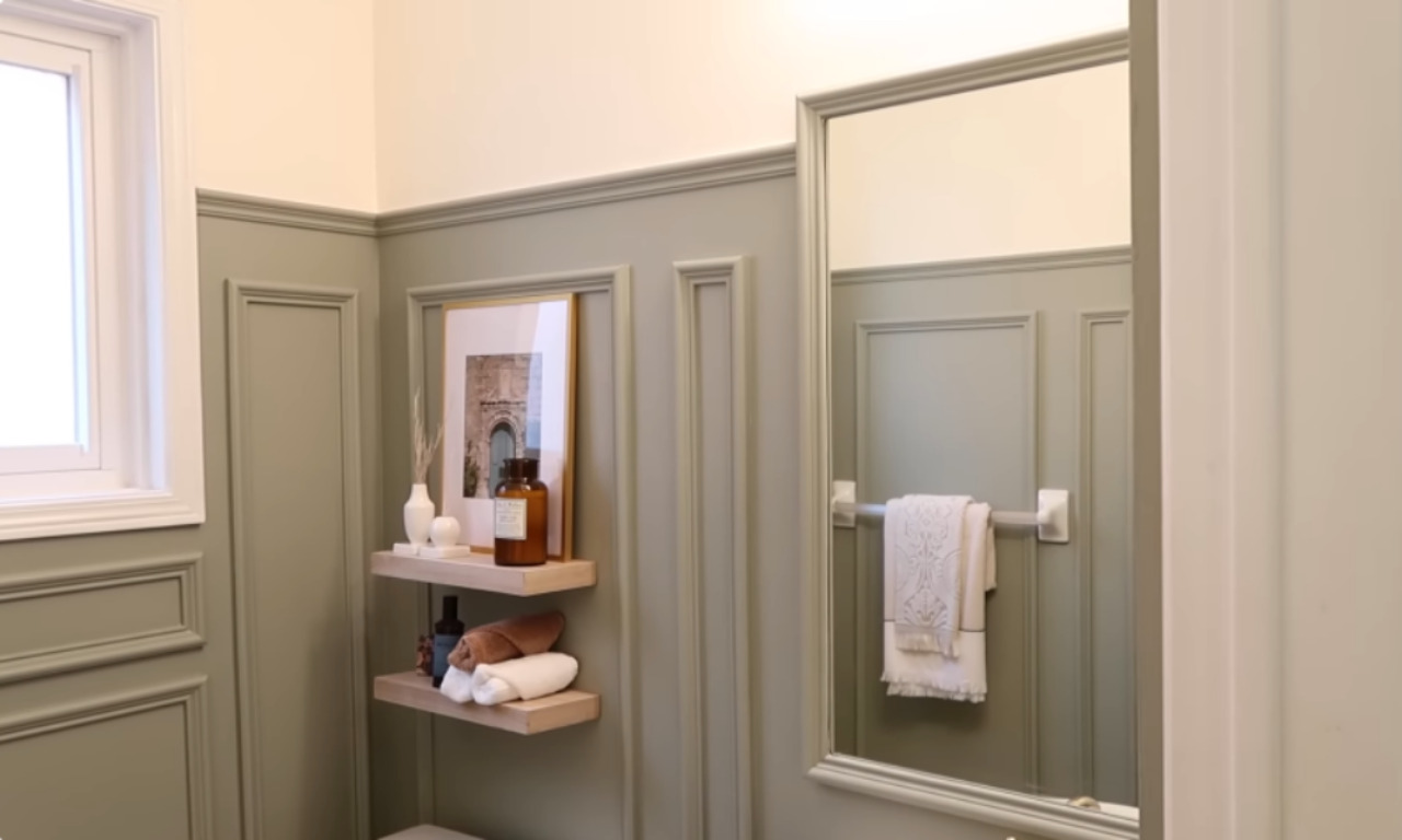 A stylish bathroom wall painted in a muted sage green with decorative wainscoting. Two light-wood floating shelves are installed on the wall, holding decorative items and neatly folded towels. A tall mirror hangs to the right.