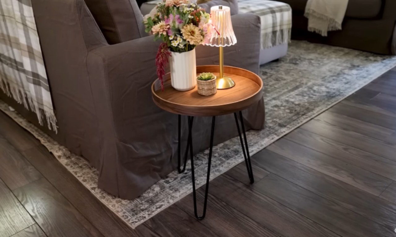 A small, round side table with a dark wood top and black hairpin legs. It holds a white vase of flowers, a small succulent, and a bedside lamp with a pink pleated shade. The table sits on a patterned rug in front of a gray slipcovered sofa.
