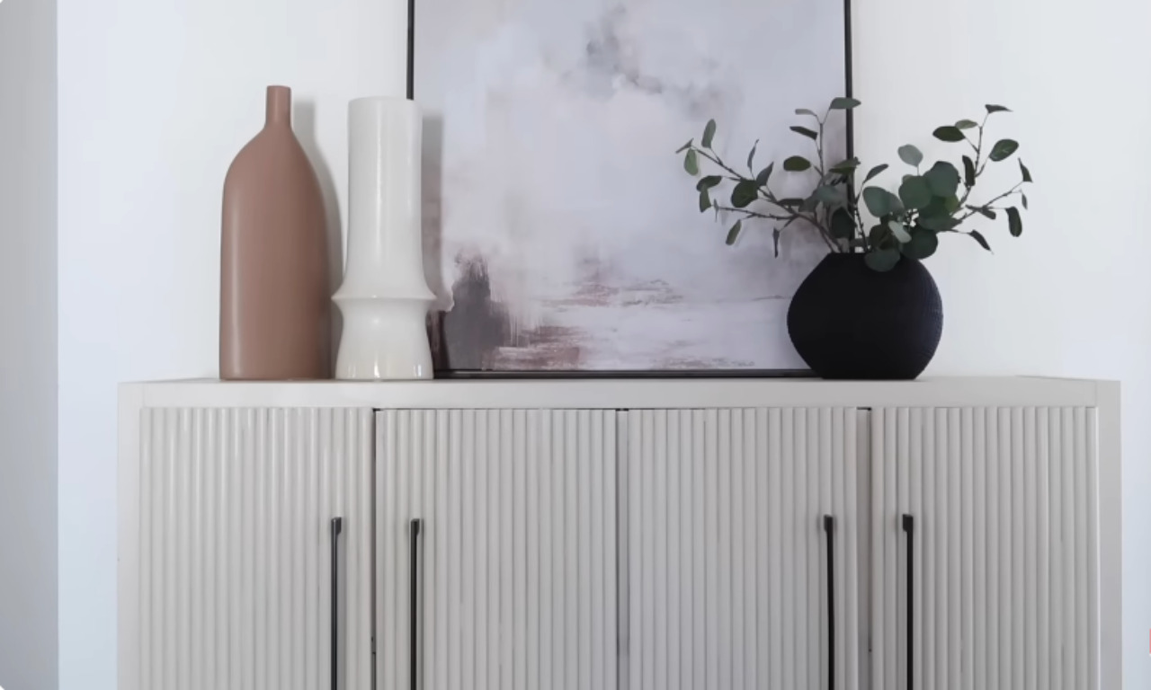 A close-up view of the top of a finished, modern sideboard. The cabinet doors have a textured, fluted or reeded design, painted a pale off-white or beige. The top of the cabinet is styled with two ceramic vases (one terracotta/pink, one tall white) and a black vase holding a green eucalyptus-like plant, set in front of a framed abstract painting with soft beige and gray tones.