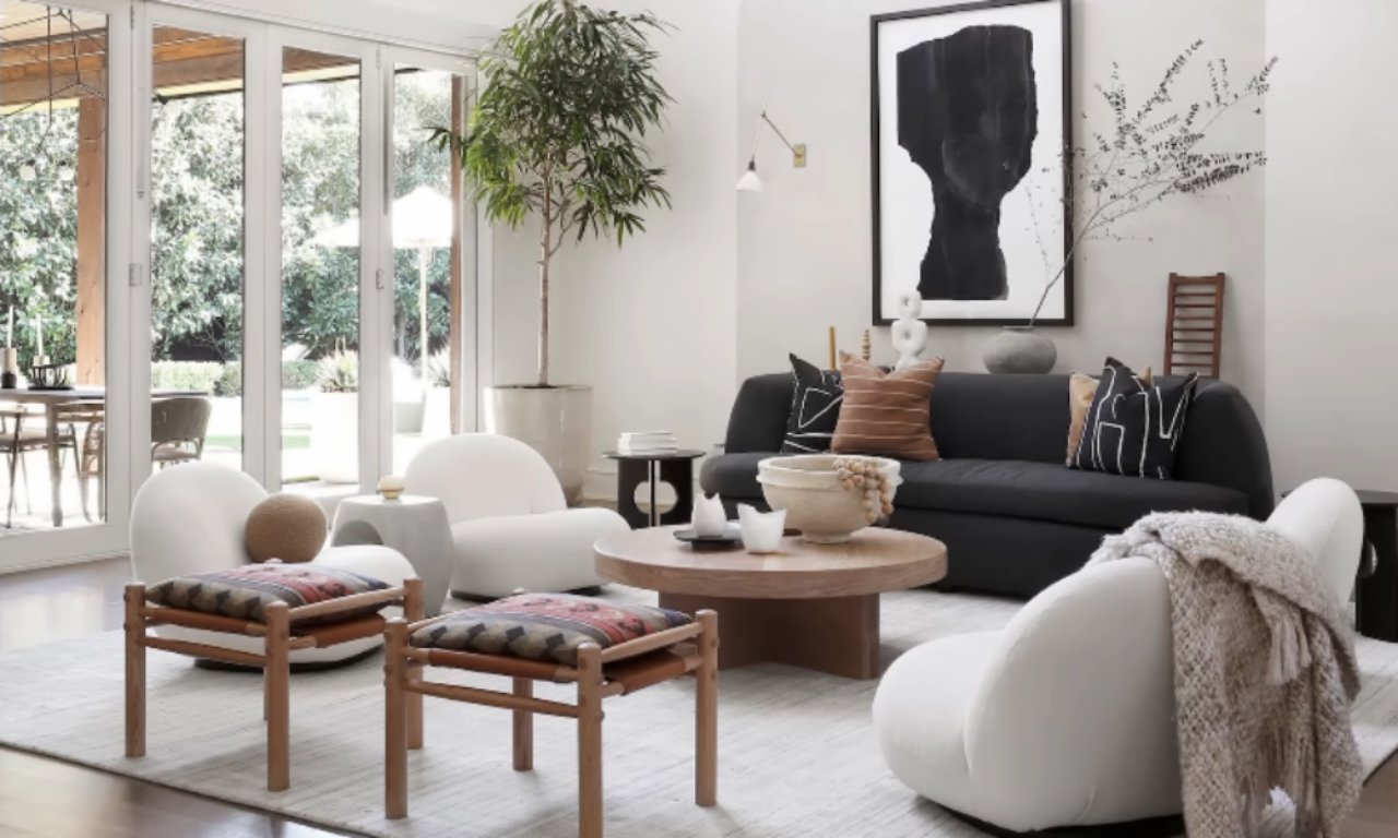 A bright, modern living room with glass doors opening to the outdoors. The room features a dark gray sofa with patterned throw pillows, two white, curved low armchairs, and a round wooden coffee table. Two low wooden stools with patterned, colorful fabric seats are in the foreground. A large black and white abstract art print hangs on the white wall.