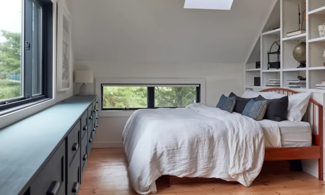 A bright, modern bedroom with sloped ceilings and built-in shelving on the right wall filled with books and decor. The white bed with a wooden frame is covered in a loose, white linen duvet and has dark blue and patterned accent pillows. A long, dark teal or navy blue dresser/cabinet runs along the left wall under two windows. A large window on the left has a black frame, and a smaller, horizontal window is positioned behind the bed, looking out onto a wooded area.
